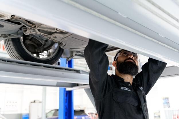 A certified mechanic wearing safety goggles working underneath an electric vehicle that is elevated in a vehicle repair centre.